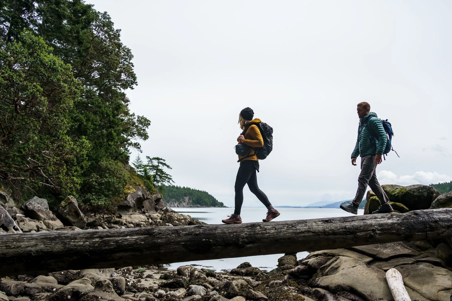 two individuals walking on a log balanced on rocks beside the ocean